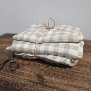 Op de afbeelding: Een stapel gevouwen, beige en wit geruite stof, vastgebonden met touw. Een antieke schaar ligt op een houten oppervlak naast de stof. De stof heeft een klassiek ruitpatroon.