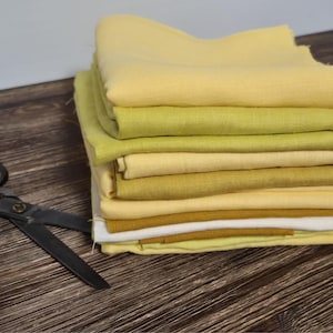 Può includere: Una pila di tessuto piegato in varie tonalità di giallo e verde, con un paio di forbici nere su una superficie di legno scuro. I tessuti variano dal giallo chiaro al verde scuro.