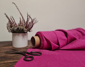 May include: A roll of bright pink fabric is laid out on a wooden table. A pair of scissors is lying on the fabric. A small white pot with dried pink flowers is in the background.