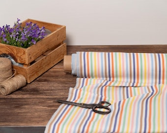 Può includere: Una cassa di legno piena di fiori viola, un rotolo di tessuto a righe, spago e forbici vintage poggiano su una superficie di legno. Il tessuto presenta strisce verticali in vari colori, tra cui blu, arancione e giallo.