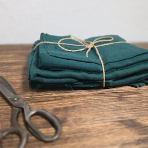 Op de afbeelding: Een stapel teal linnen servetten gebonden met touw. De servetten zijn gevouwen en gerangschikt op een houten oppervlak.