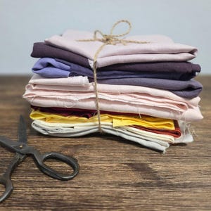 May include: A bundle of fabric squares in various colours, secured with twine. The colours include pinks, purples, yellows, and grey. A pair of vintage scissors lies on a wooden surface. Suitable for craft projects.