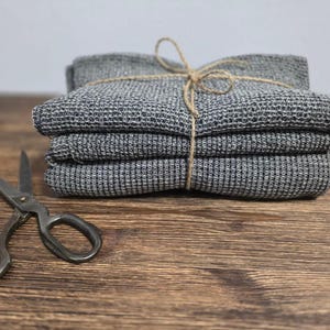 Op de afbeelding: Een stapel van drie grijze geweven handdoeken, vastgebonden met touw, naast een vintage schaar. De handdoeken hebben een getextureerde uitstraling en zijn netjes opgevouwen. De schaar ligt open op een houten oppervlak.
