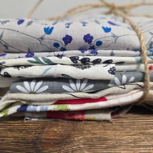 Könnte beinhalten: Ein Stapel gefalteter Stoffe mit floralen Mustern, mit Bindfaden gebunden. Die Stoffe weisen verschiedene Designs auf, darunter blaue Blumen, grüne Blätter und weiße Gänseblümchen. Die Stoffe liegen auf einer Holzoberfläche.