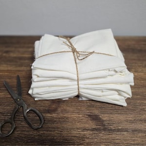 May include: A stack of white cotton tea towels tied together with twine, sitting on a wooden surface. A pair of antique scissors are lying next to the towels.