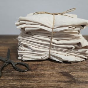 Op de afbeelding: Een stapel crèmekleurige vierkante stoffen, die met touw aan elkaar zijn gebonden. De stof is netjes gevouwen en lijkt van een natuurlijk materiaal te zijn. Een zwart schaar ligt op een houten oppervlak.