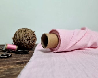 May include: A roll of pink fabric is lying on a wooden surface. A ball of brown yarn and a reel of pink thread are also on the surface. A pair of scissors is lying next to the reel of thread.