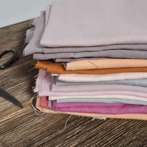 Könnte beinhalten: Ein Stapel farbiger Stoffmuster in Grau-, Orange-, Rosa- und Lilatönen. Eine antike Schere liegt auf einer Holzoberfläche neben dem Stoff. Die Stoffe scheinen aus Leinen oder Baumwolle zu sein.
