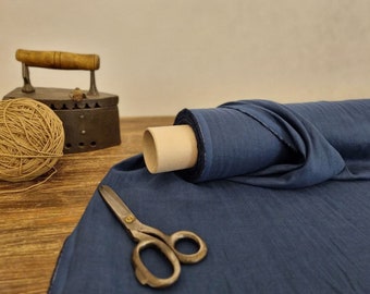 May include: A roll of navy blue fabric sits on a wooden surface, alongside vintage scissors, a ball of twine, and an antique iron. The fabric is partially unrolled, revealing its texture and color. The scene evokes a sense of traditional craftsmanship.
