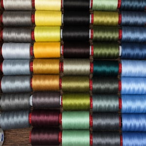 Op de afbeelding: Een verzameling naaigarenspoelen gerangschikt in een rasterpatroon. De spoelen hebben een grote verscheidenheid aan effen kleuren, waaronder tinten bruin, grijs, geel, zwart, groen en blauw, elk gewikkeld op een kern met rode eindkappen.