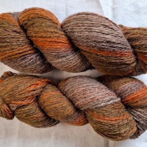 Puede incluir: Dos madejas de hilo grueso de varias hebras con una paleta de colores variados de marrón tierra, verde oliva intenso y naranja quemado vibrante. Las fibras tienen una textura ligeramente difusa y rústica con una torsión visible.