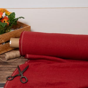 Könnte beinhalten: Eine Rolle roter Stoff mit einem ausgerollten Abschnitt, der die Textur zeigt. Eine Holzkiste mit orangefarbenen Gerbera-Gänseblümchen, Bindfaden und einer Schere liegt auf einer Holzoberfläche.