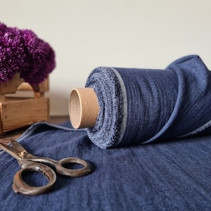 May include: A roll of dark blue denim fabric with a cardboard core sits on a matching fabric surface. Vintage scissors are open on the fabric. A wooden crate holds purple flowers. Spools of thread are in the background.