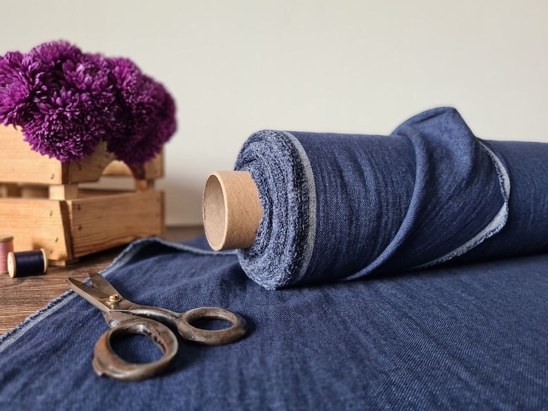 May include: A roll of dark blue denim fabric with a cardboard core sits on a matching fabric surface. Vintage scissors are open on the fabric. A wooden crate holds purple flowers. Spools of thread are in the background.