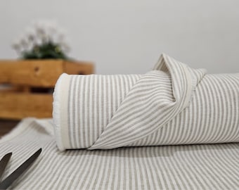 May include: A roll of beige and white striped fabric. The fabric is rolled up, with a section unrolled and draped over the roll. A pair of scissors rests on the fabric. The fabric has a natural, textured appearance, suitable for home decor or sewing projects.