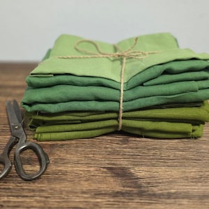 Könnte beinhalten: Ein Stapel grüner Stoffquadrate in verschiedenen Schattierungen, mit Bindfaden gebunden. Eine antike Schere liegt auf einer Holzoberfläche neben dem Stoff. Die Stofffarben reichen von hell- bis dunkelgrün.