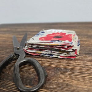May include: A pile of small, square fabric pieces with a floral design, including red poppies and blue details, on a neutral backdrop. A pair of vintage scissors with dark metal handles rests beside the fabric. The items are on a wooden surface.