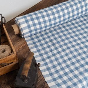 Puede incluir: Un rollo de tela a cuadros azul y blanco descansa sobre una superficie de madera, junto a una caja de madera llena de hilo y tijeras. También hay una plancha antigua. La tela está desenrollada, mostrando el patrón de cuadros.