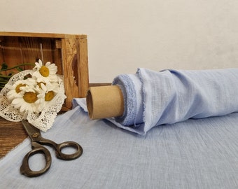 May include: A roll of light blue fabric with a textured weave. A bouquet of white daisies with yellow centers and a pair of vintage scissors are next to the fabric. A wooden crate is in the background.