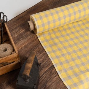 Op de afbeelding: Een rol geel en grijs geruite stof ligt op een houten oppervlak. Een houten kist bevat strengen touw en een schaar. Een antieke strijkijzer is ook aanwezig. Geschikt voor naaiprojecten.