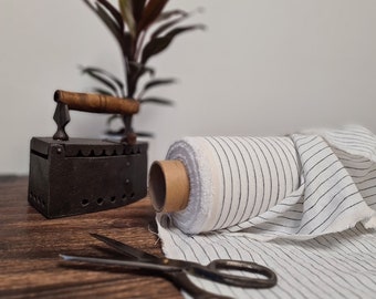 May include: A vintage flat iron with a wooden handle sits near a roll of striped fabric and a pair of scissors. The fabric is white with thin black vertical stripes. The iron is dark gray and the scissors are silver.