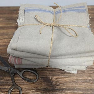 Puede incluir: Un montón de tela de lino gris claro doblada, atada con hilo. La tela tiene rayas azules y rojas. Un par de tijeras vintage con mangos negros se encuentra sobre una superficie de madera.