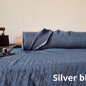 May include: A roll of silver-blue fabric with a textured appearance. The fabric is displayed on a wooden surface, alongside a vintage iron, scissors, and thread. The text "Silver blue" is visible.