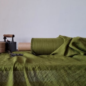 May include: A roll of olive green fabric with a textured, semi-sheer weave. The fabric is partially unrolled, draping across the surface. Nearby, a vintage black metal iron, a spool of brown twine, and a pair of black scissors are visible.