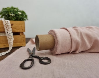 May include: A roll of blush pink fabric sits on a wooden surface, with a pair of vintage scissors and a length of lace nearby. The fabric is unrolled, revealing its texture. A small wooden crate with greenery is in the background.