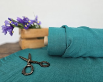 Op de afbeelding: Een rol teal-kleurige stof ligt op een houten oppervlak, met een vintage schaar en een stuk stof uitgespreid. Paarse irissen staan in een houten krat op de achtergrond.