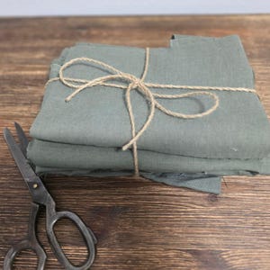 May include: A stack of sage green fabric squares tied with twine, next to a pair of vintage scissors. The fabric is neatly folded and appears to be linen or a similar material. The scissors are open, suggesting they are ready for use.