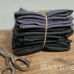 Può includere: Un mucchio di quadrati di tessuto nero e grigio legati insieme con uno spago. Il tessuto è piegato ordinatamente e i bordi sono sfilacciati. Il tessuto è su una superficie di legno. Toni neri.