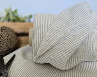 May include: A roll of beige and white striped fabric, likely linen or cotton, is the focal point. The fabric is folded and draped, showcasing the pattern. A ball of twine and scissors are in the foreground, suggesting a crafting or sewing theme.