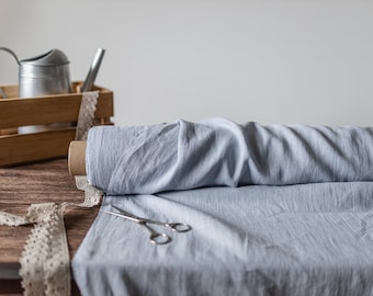 May include: A roll of light blue fabric with a textured surface lies on a wooden surface next to a pair of silver metal scissors. A wooden crate containing a silver metal watering can and a strip of beige lace trim sits nearby.