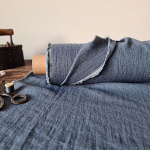 Puede incluir: Un rollo de tela azul con un borde sin rematar se encuentra sobre una superficie de madera. Tambi&eacute;n hay una plancha antigua, tijeras e hilo, lo que sugiere un tema de costura o textil. La tela parece ser de peso medio.
