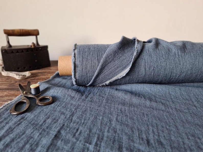 Puede incluir: Un rollo de tela azul con un borde sin rematar se encuentra sobre una superficie de madera. Tambi&eacute;n hay una plancha antigua, tijeras e hilo, lo que sugiere un tema de costura o textil. La tela parece ser de peso medio.