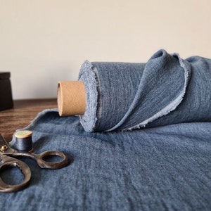 Puede incluir: Un rollo de tela azul con un borde sin rematar se encuentra sobre una superficie de madera. Tambi&eacute;n hay una plancha antigua, unas tijeras y una bobina de hilo, lo que sugiere un tema de costura o artesan&iacute;a. La tela es de color azul medio.