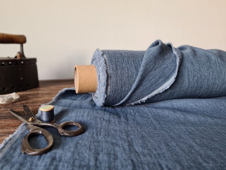 Puede incluir: Un rollo de tela azul con un borde sin rematar se encuentra sobre una superficie de madera. Tambi&eacute;n hay una plancha antigua, unas tijeras y una bobina de hilo, lo que sugiere un tema de costura o artesan&iacute;a. La tela es de color azul medio.