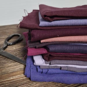 Op de afbeelding: Een stapel gekleurde stofstalen in tinten paars, roze en blauw, met een vintage schaar op een houten oppervlak. De stof lijkt linnen of een vergelijkbaar materiaal te zijn, met zichtbare textuur.