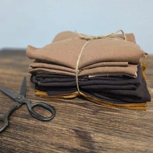 May include: A stack of brown and black fabric squares tied together with twine. The fabric is folded neatly and the twine is tied in a bow. A pair of black scissors are lying on a wooden surface.