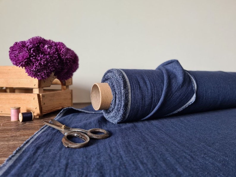May include: A roll of dark blue fabric sits next to a pair of vintage scissors. A wooden crate holds a cluster of purple flowers. Two spools of thread, one pink and one blue, are also visible.