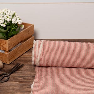 May include: A roll of red and white striped fabric sits next to a wooden crate holding a potted plant with white flowers. A spool of twine and vintage scissors are also on the wooden surface.