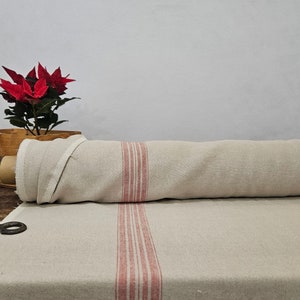 Puede incluir: Un rollo de tela de color natural con rayas rojas, parcialmente desenrollado sobre una superficie de madera. También se ve una caja de madera con una planta de hojas rojas y tijeras, lo que sugiere un tema artesanal o de decoración del hogar.