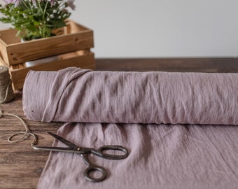Puede incluir: Un rollo de tela malva descansa sobre una mesa de madera, con unas tijeras antiguas y un carrete de hilo cerca. Una caja de madera con una planta en maceta está en el fondo. La tela parece ser lino o un material similar.
