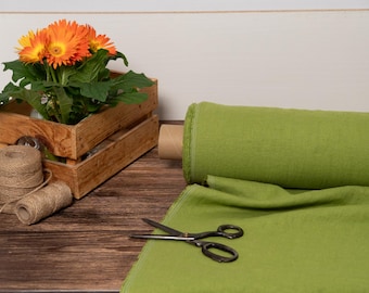 May include: A still life composition with a wooden crate holding orange gerbera daisies, a roll of green fabric, twine, and scissors. The fabric is unrolled, and the scissors are open. The scene is set on a wooden surface.