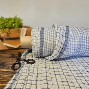 Könnte beinhalten: Eine Rolle blau-weiß karierter Stoff liegt teilweise auf einer Holzoberfläche ausgerollt. Eine Schere und eine Holzkiste mit Grün und Spitze befinden sich im Hintergrund. Der Stoff eignet sich für Heimdekoration und Bastelprojekte.