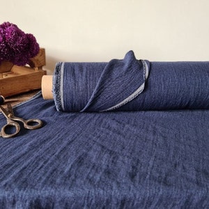 May include: A roll of navy blue fabric with a textured appearance. The fabric is unrolled on a wooden surface, with a pair of vintage scissors and a small wooden crate with purple flowers in the background. The fabric is on a cardboard roll.