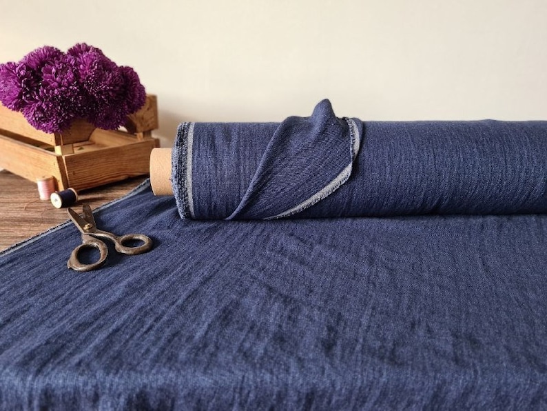 May include: A roll of navy blue fabric with a textured appearance. The fabric is unrolled on a wooden surface, with a pair of vintage scissors and a small wooden crate with purple flowers in the background. The fabric is on a cardboard roll.