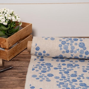 Pode incluir: Um rolo de tecido bege com um padrão floral azul é exibido, ao lado de uma caixa de madeira com uma planta com flores brancas. Barbante marrom e tesouras também são visíveis, sugerindo um tema de artesanato ou decoração.
