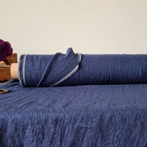 May include: A roll of dark blue fabric with a textured appearance. The fabric is partially unrolled, revealing a lighter edge. A wooden crate with purple flowers and a pair of scissors are in the background.